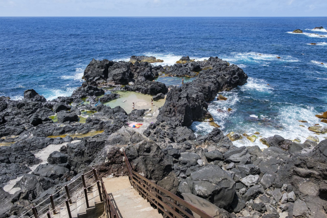 Natural seawater pools at Poceir&otilde;es on Graciosa Island, surrounded by rugged black volcanic rock and the open Atlantic Ocean. Wooden stairs descend through the basalt formations to the sheltered swimming area.