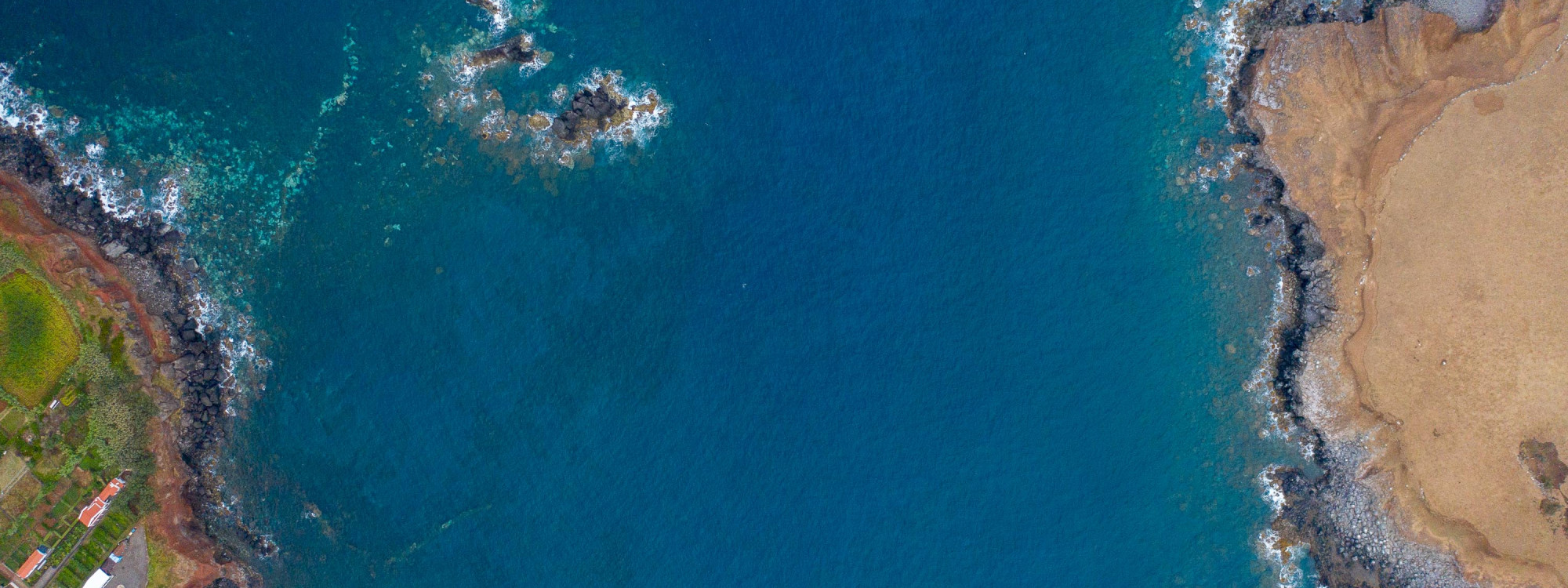 Top-down aerial view of dark basalt formations surrounded by clear Atlantic water along the volcanic coast of São Jorge Island.