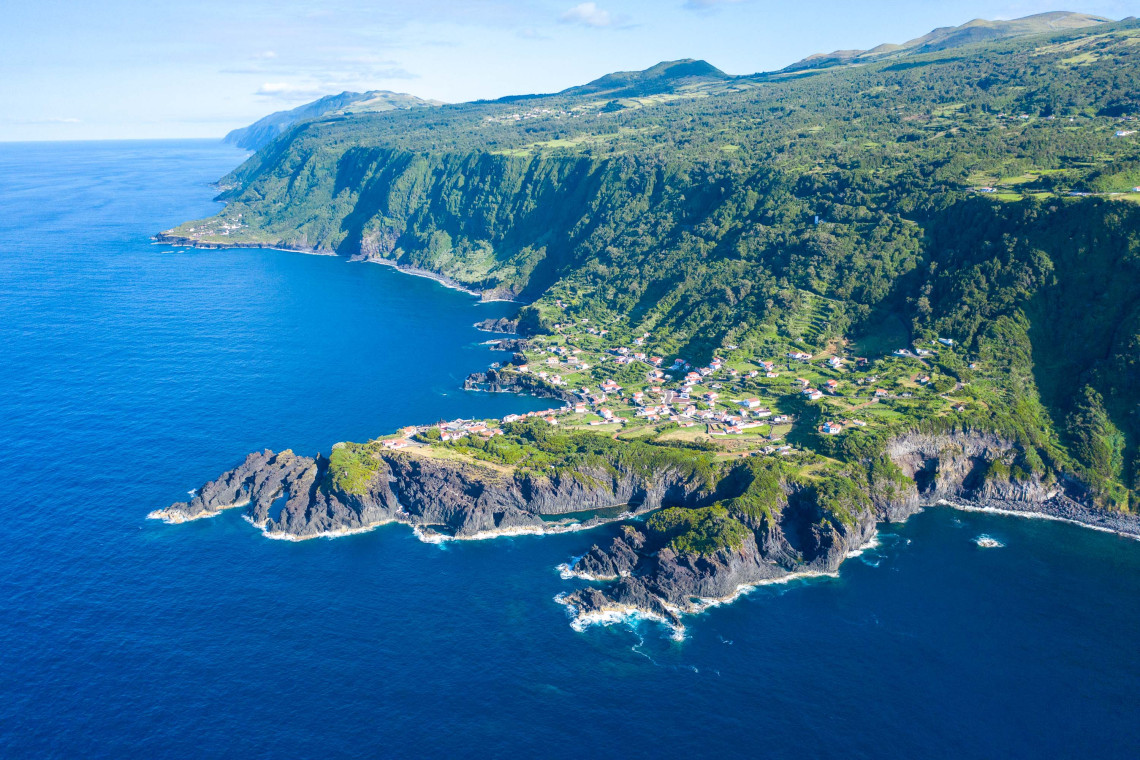 Aerial view of Faj&atilde; do Ouvidor on S&atilde;o Jorge Island, showing its village spread across a volcanic platform beneath steep green cliffs and beside the Atlantic Ocean.