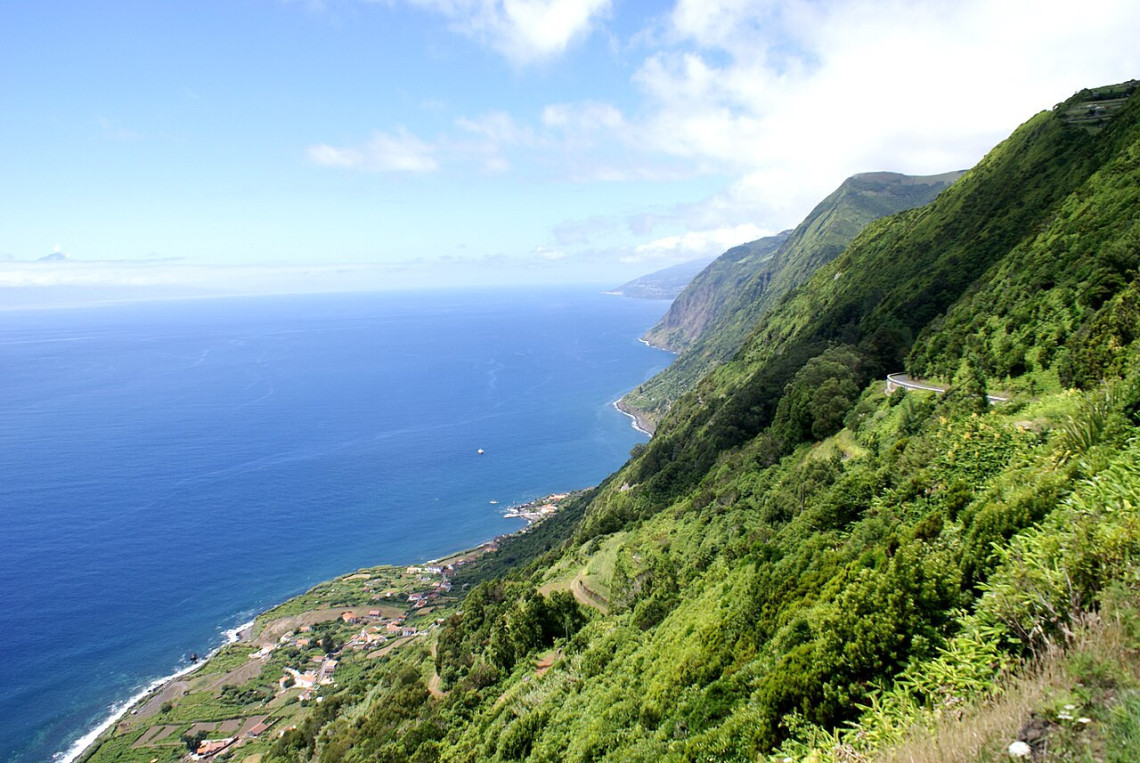Elevated view along the southern coast of S&atilde;o Jorge Island toward Faj&atilde; dos Vimes, with steep green cliffs descending to farmland and houses beside the Atlantic.