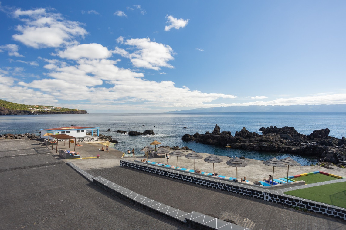 Traveler relaxing at the volcanic pools of Po&ccedil;a dos Frades in Velas, S&atilde;o Jorge, with dark basalt formations and Mount Pico visible across the Atlantic channel.
