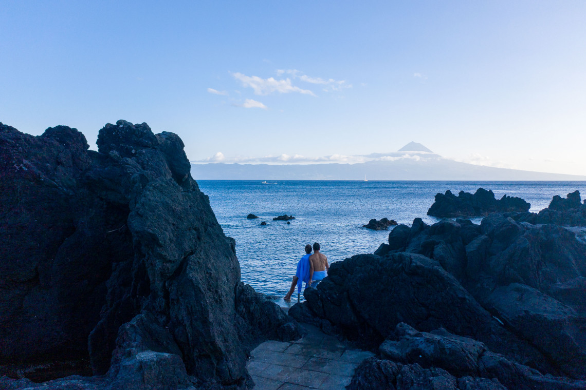 Visitors sitting among dark volcanic rocks at Po&ccedil;a dos Frades in Velas, S&atilde;o Jorge, with the Atlantic Ocean and Mount Pico visible in the distance.