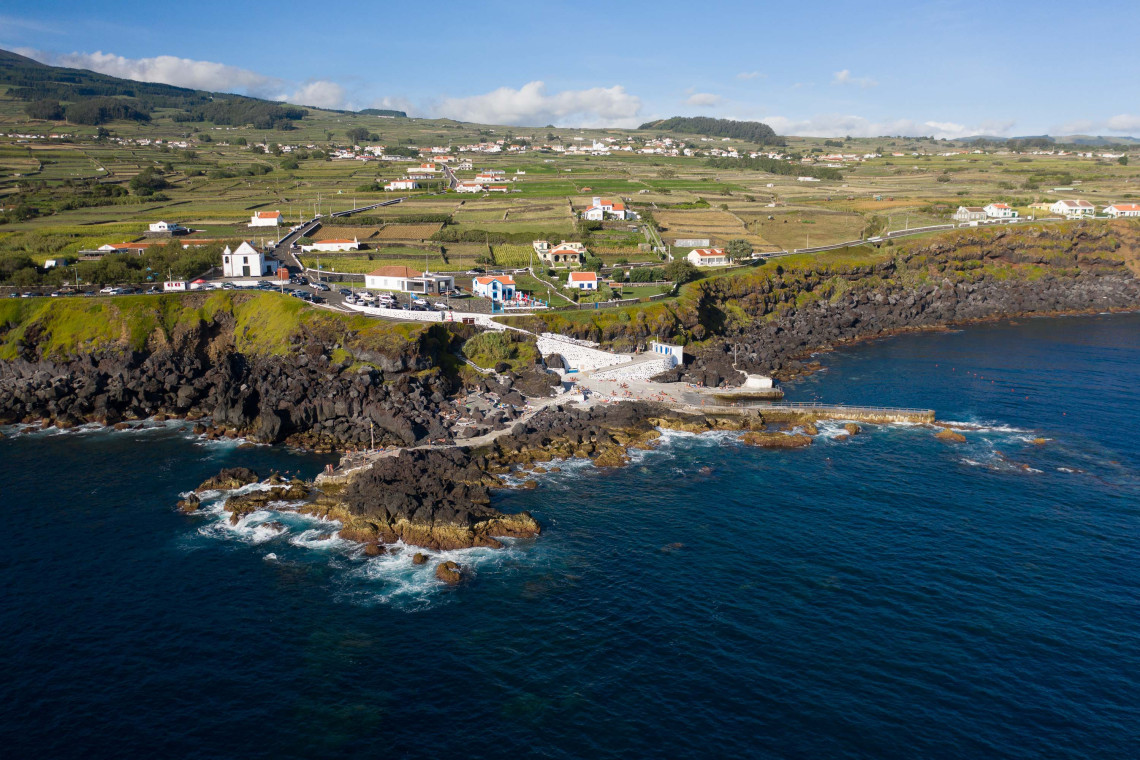 Aerial view of Porto das Cinco Ribeiras on Terceira Island, showing the volcanic coastline, natural swimming area and green agricultural fields above the Atlantic Ocean.