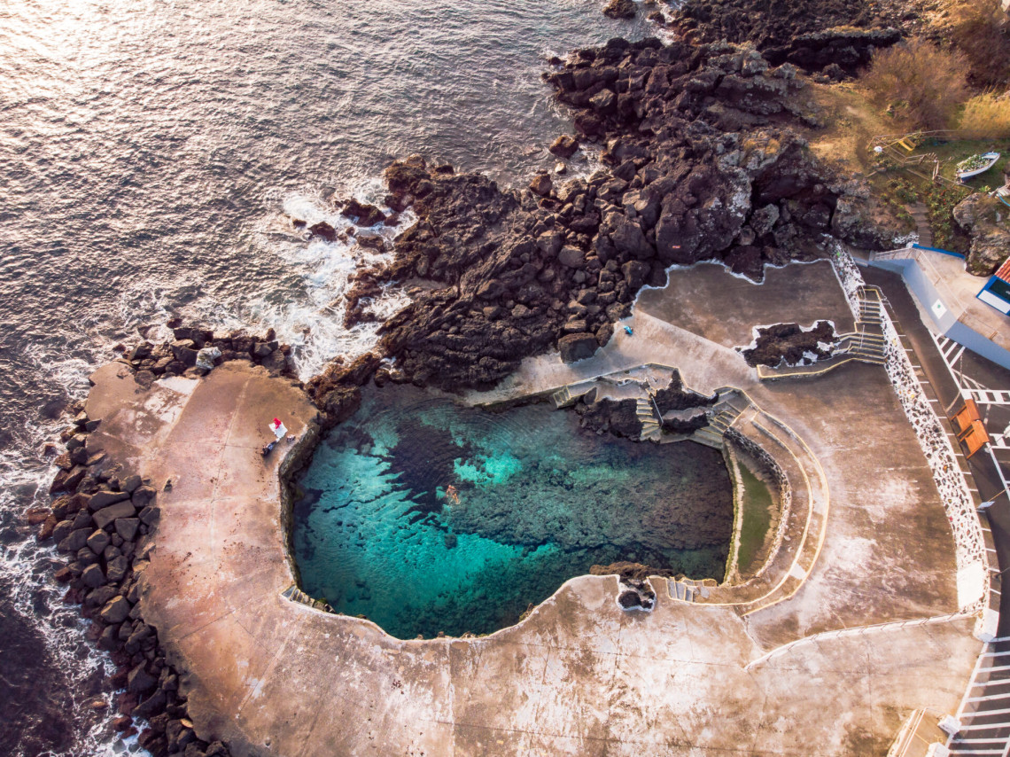Aerial view of the Piscina Natural do Refugo in Porto Judeu on Terceira Island, with clear seawater enclosed by volcanic rocks and concrete bathing platforms.