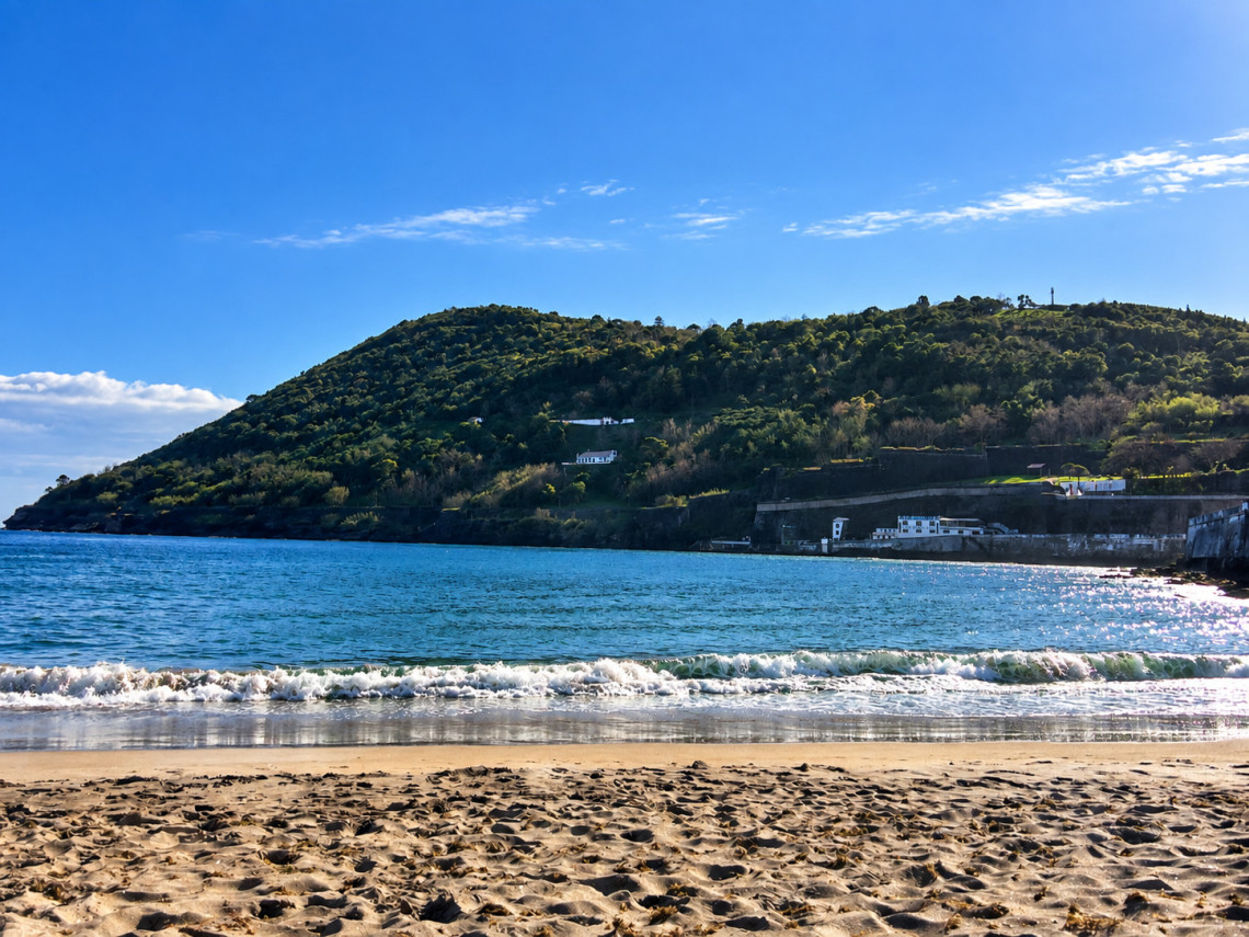 View from Prainha in Angra do Hero&iacute;smo across the Atlantic bay toward the green slopes of Monte Brasil, with gentle waves reaching the sandy beach.