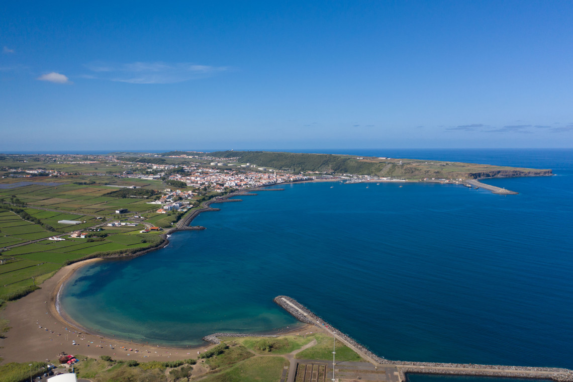 Wide aerial view of Praia da Vit&oacute;ria on Terceira Island, showing the curved sandy beach, marina, coastal city and expansive blue Atlantic bay.