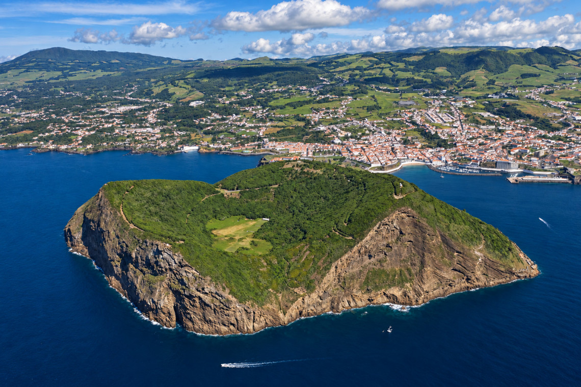 Aerial view of Monte Brasil, the volcanic peninsula beside Angra do Hero&iacute;smo on Terceira Island, with its forested crater, steep Atlantic cliffs and the city extending around the bay in the background.