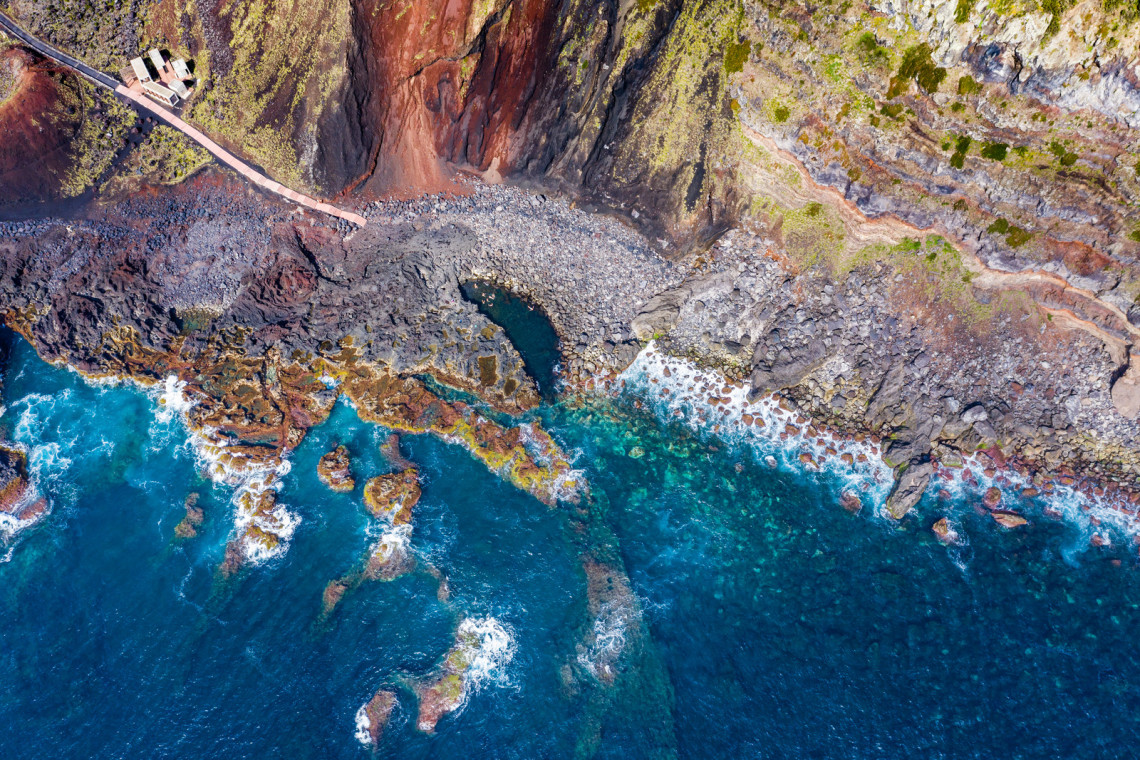 Aerial view of the natural swimming pool at Ponta da Ferraria in Ginetes, S&atilde;o Miguel Island. A footpath crosses the dark volcanic shoreline beside clear Atlantic water and layered coastal cliffs.