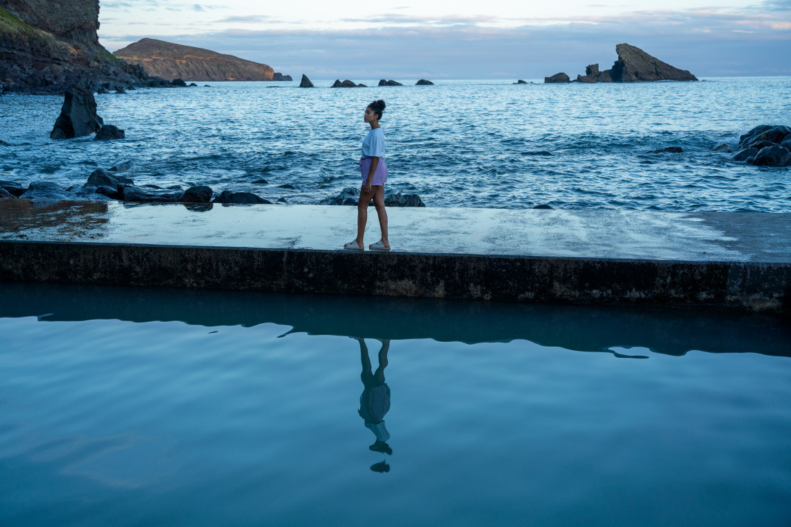 A person walks beside the outdoor thermal pool at Termas do Carapacho on Graciosa Island in the Azores, Portugal. Volcanic rock formations and the Atlantic Ocean extend across the background in the evening light.