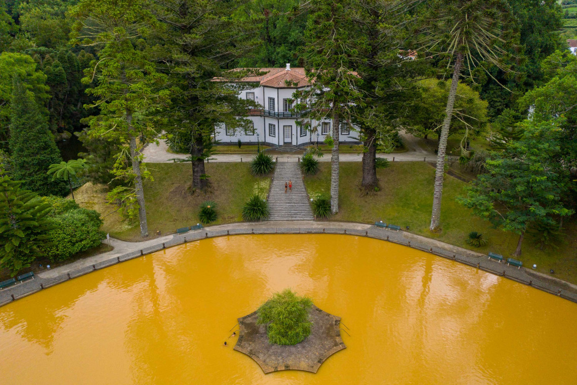 Aerial view of Terra Nostra Park in Furnas, showing the large ochre thermal pool, central planted island and historic garden building. Mature trees and subtropical vegetation surround the bathing area on S&atilde;o Miguel Island.