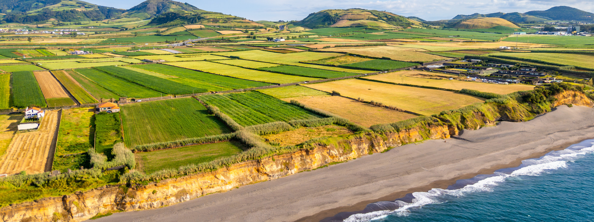 Aerial view of Areal de Santa Bárbara on São Miguel Island, showing the long volcanic sand beach below a patchwork of green and golden agricultural fields.
