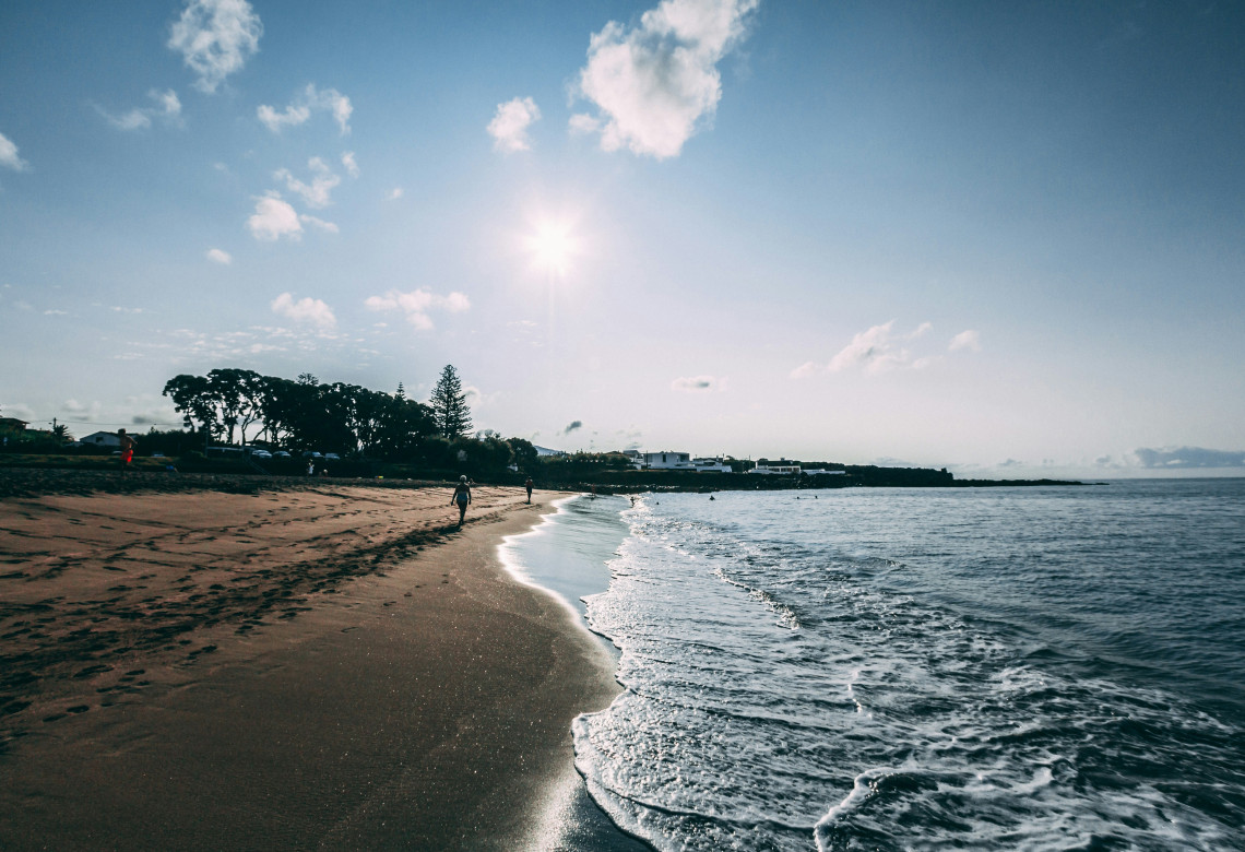 Praia do P&oacute;pulo in Ponta Delgada on S&atilde;o Miguel Island, with dark volcanic sand, gentle Atlantic waves and people walking along the shoreline under a bright sky.