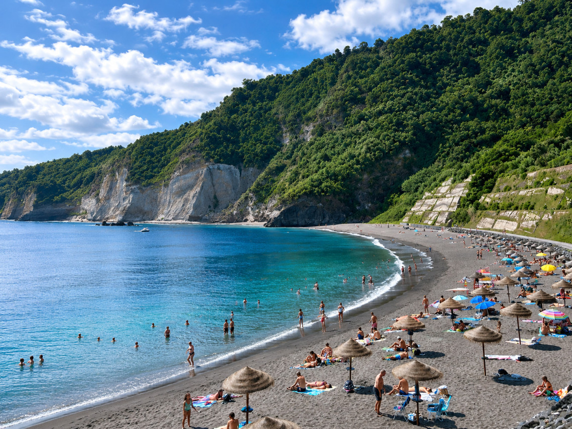 Swimmers and beachgoers at Praia do Fogo in Ribeira Quente, with dark volcanic sand and clear Atlantic water beneath the steep green cliffs of S&atilde;o Miguel Island.