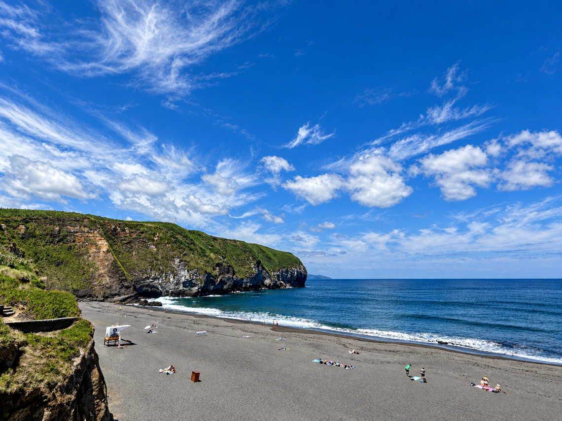 Wide volcanic sand beach at Areal de Santa B&aacute;rbara on S&atilde;o Miguel Island, with beachgoers beside the Atlantic Ocean and green cliffs framing the coastline.