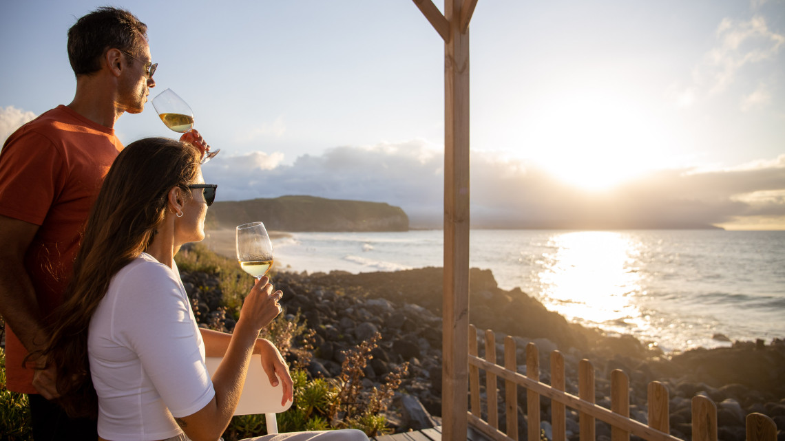 Couple enjoying glasses of wine overlooking Areal de Santa B&aacute;rbara in Ribeira Grande on S&atilde;o Miguel Island, with the Atlantic Ocean and the long volcanic-sand coastline illuminated by the setting sun.