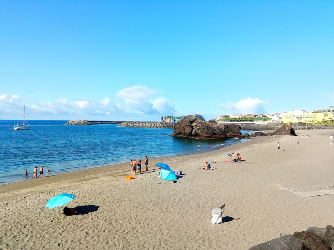 Praia da Vinha da Areia in Vila Franca do Campo, with beachgoers on the sand, calm blue Atlantic water and volcanic rock formations near the marina.