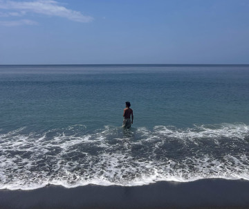 Person standing in the Atlantic Ocean along a dark volcanic sand beach on São Miguel Island in the Azores, with gentle waves reaching the shoreline.