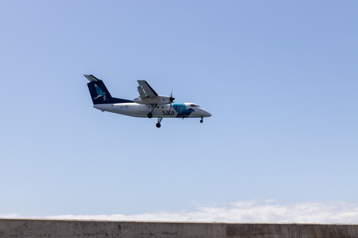An aircraft stands on the runway at Corvo Airport in Vila do Corvo, Azores, Portugal. White houses with red-tiled roofs and steep volcanic slopes rise immediately behind the small island airport.