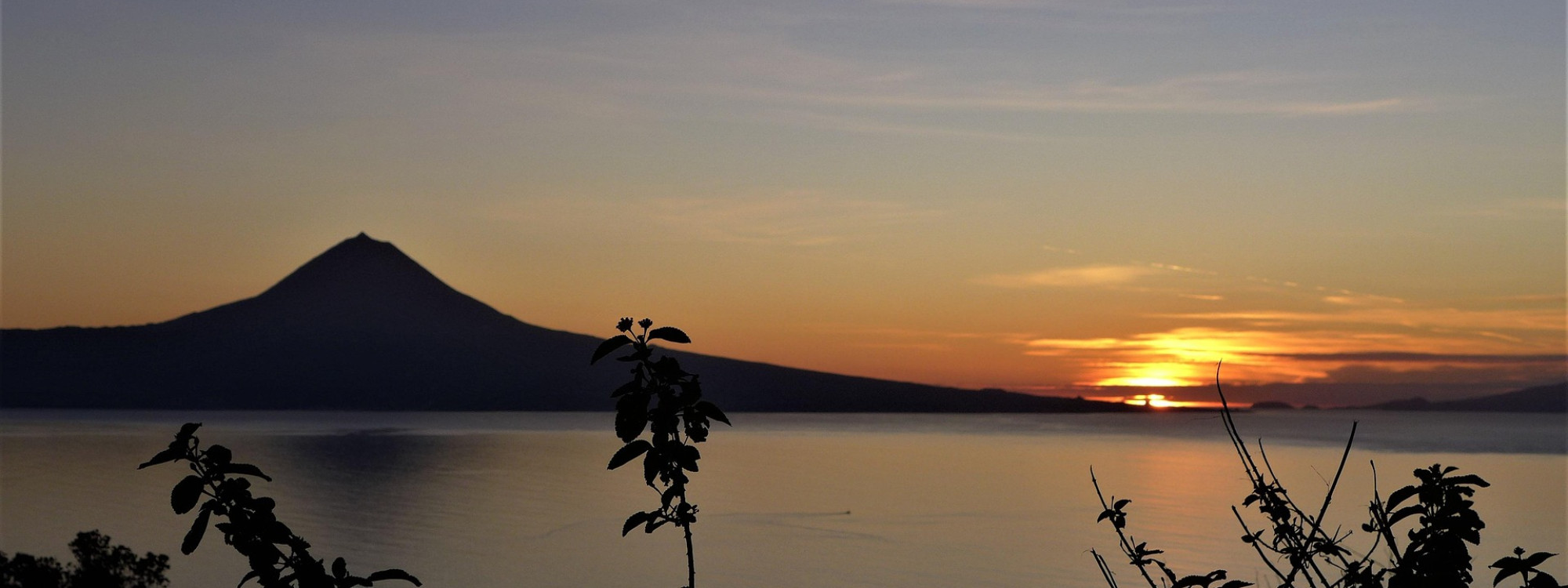 Sunset view from Faial Island toward Mount Pico across the Atlantic channel, with the volcanic peak silhouetted against an orange sky.