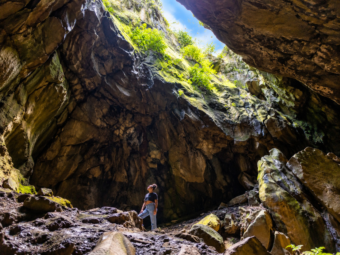 Interior view of Furna do Enxofre on Graciosa Island, showing the dramatic volcanic cave walls, natural skylight opening above, and a visitor standing inside the crater-like chamber.