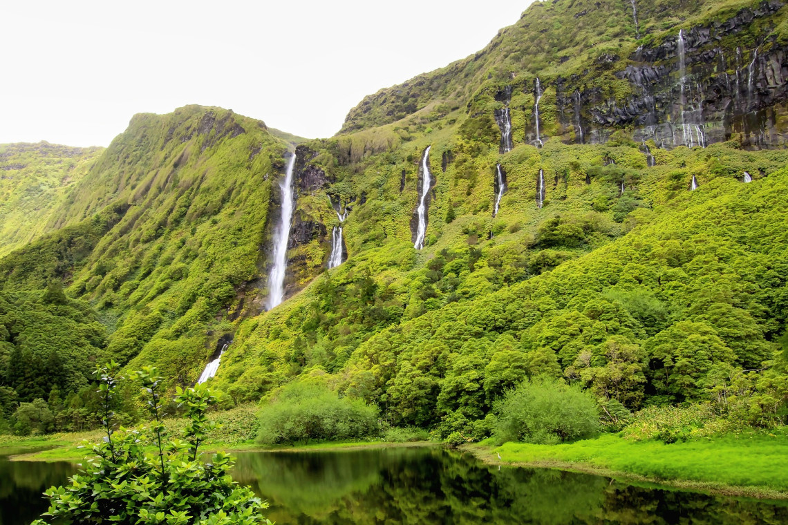 Multiple waterfalls descend the green cliffs at Po&ccedil;o da Ribeira do Ferreiro on Flores Island into a calm lagoon surrounded by dense vegetation.
