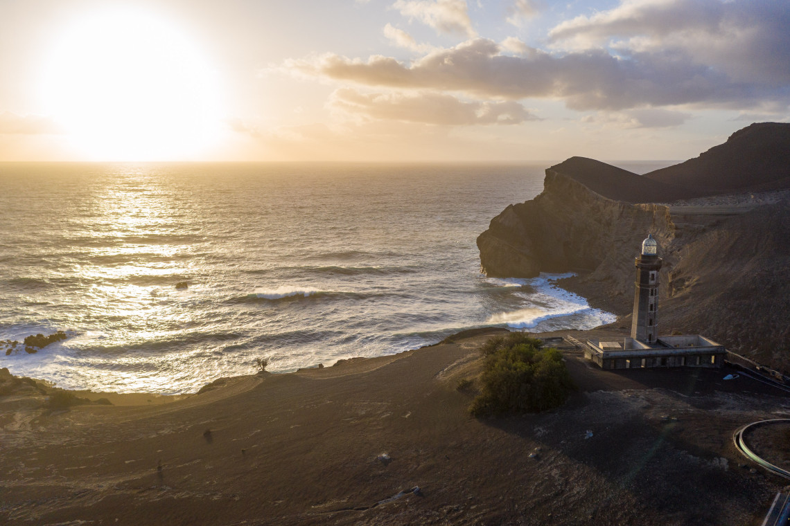 Capelinhos Lighthouse standing on the dark volcanic landscape of western Faial Island, with Atlantic waves, steep cliffs and warm sunset light in the background.