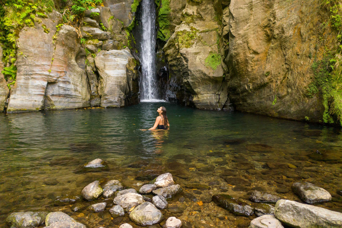 Visitor swimming in the natural pool beneath Salto do Cabrito waterfall near Ribeira Grande, surrounded by volcanic rock and dense vegetation.