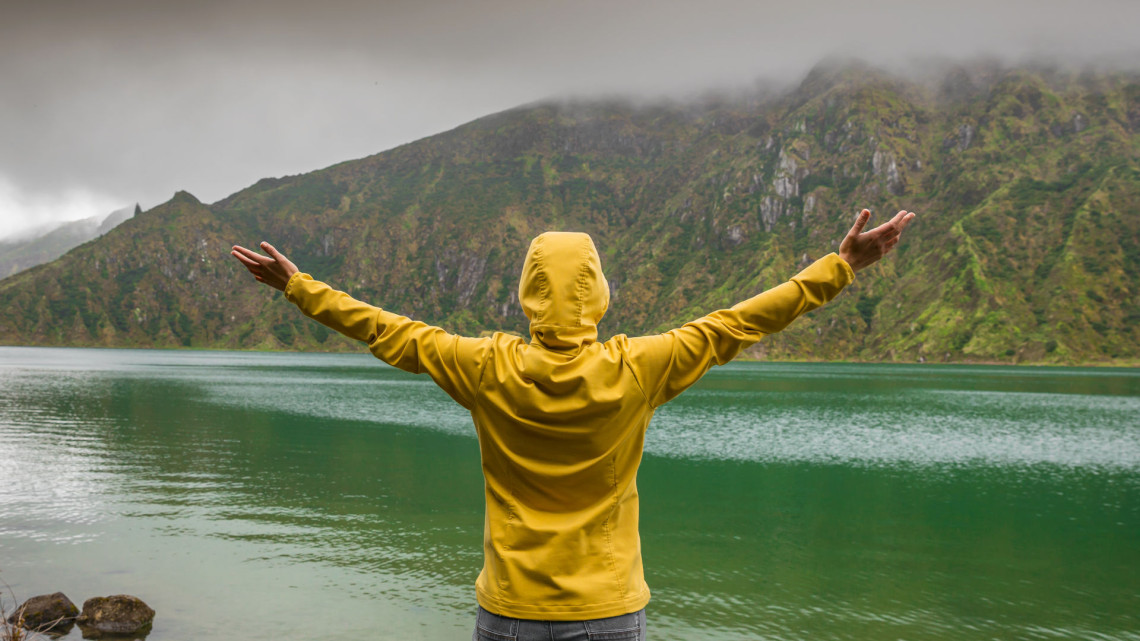 Person in a yellow raincoat with arms outstretched standing in front of the vivid green waters of Lagoa do Fogo crater lake with misty volcanic mountains on S&atilde;o Miguel Island, Azores, Portugal