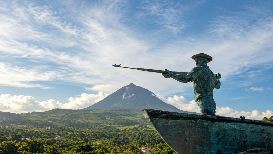 A bronze statue depicting a traditional Azorean whaler holding a harpoon at the bow of a boat, with the volcanic peak of Mount Pico rising above the green landscape in the background.