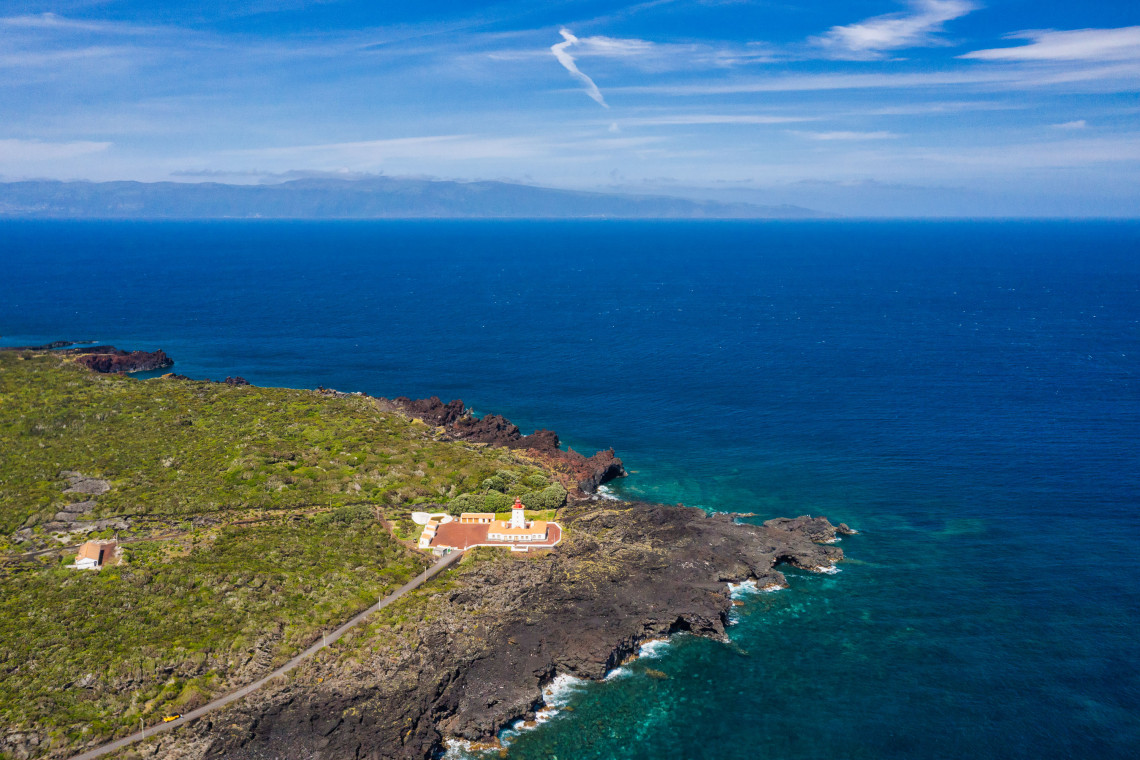 Aerial view of Ponta da Ilha Lighthouse on Pico Island, surrounded by black volcanic coastline and the blue Atlantic Ocean near Manhenha.