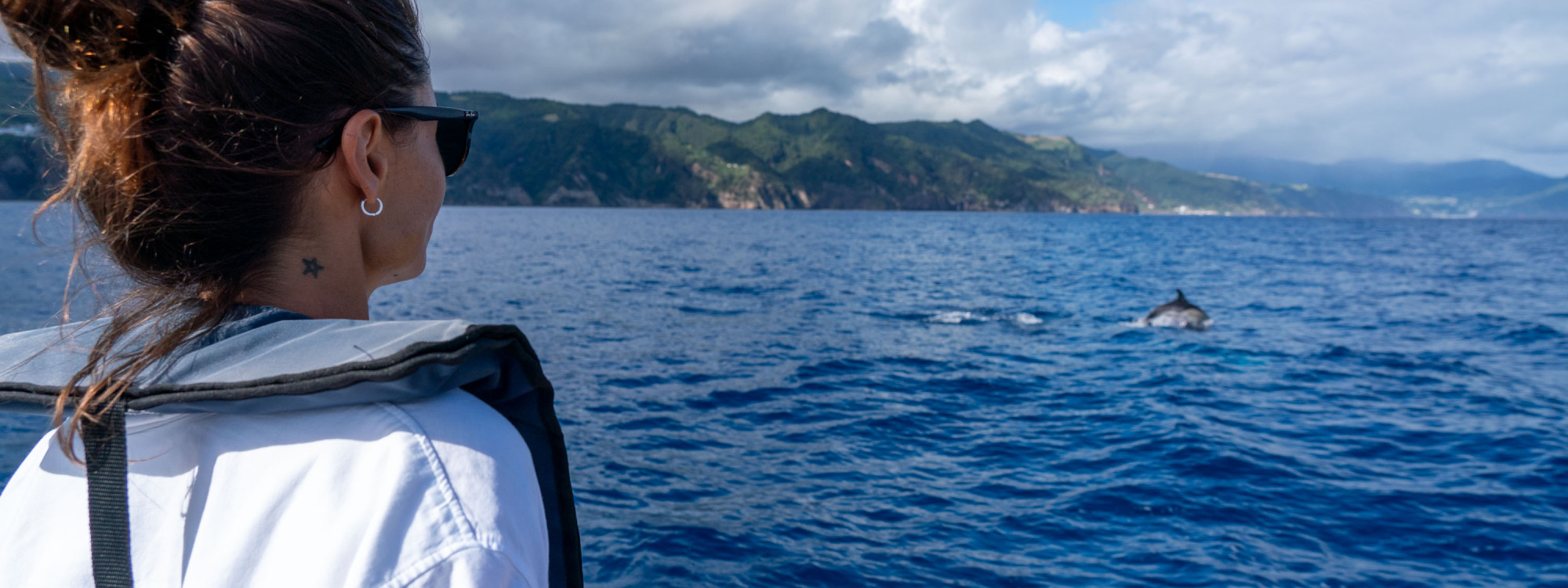 Visitor watching a dolphin surface beside a boat off the coast of São Miguel Island, with the island’s green volcanic coastline in the background.