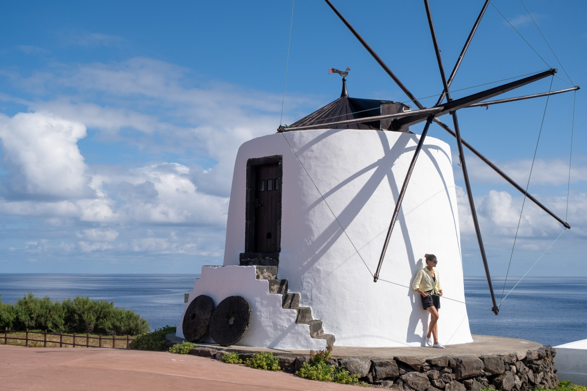 A traditional whitewashed windmill stands beside the Atlantic coast in Vila do Corvo on Corvo Island, Azores, Portugal. Its dark wooden sail structure contrasts with the white cylindrical walls and blue ocean beyond.
