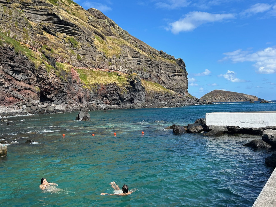 Visitors swimming in the clear seawater of the Carapacho Natural Pools on Graciosa Island, surrounded by basalt cliffs and volcanic rock formations.