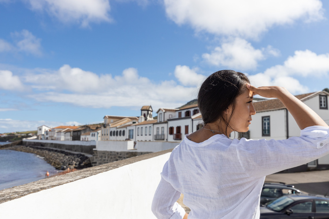 Traveler overlooking the waterfront of Vila da Praia in S&atilde;o Mateus, Graciosa, with whitewashed houses, red-tiled roofs and the Atlantic coastline behind her.
