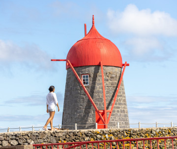 Traveler walking beside Moinho de Pedra in São Mateus on Graciosa Island, a traditional basalt windmill topped with the distinctive red wooden cap associated with the island.
