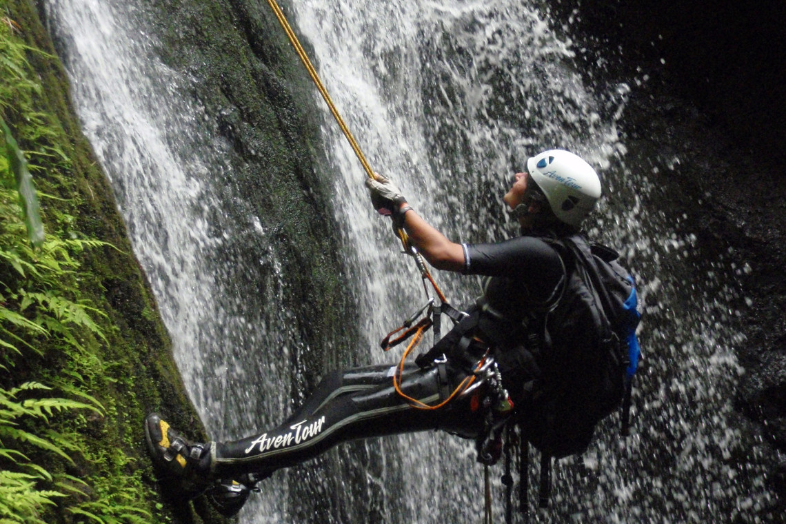 Canyoning participant rappelling beside a waterfall on S&atilde;o Jorge Island, equipped with a helmet, harness and ropes amid dense vegetation.