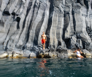 Swimmers beside the distinctive vertical basalt columns of Poça Simão Dias at Fajã do Ouvidor on São Jorge Island, surrounded by clear Atlantic water.