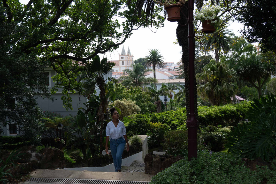Traveler walking through Jardim Duque da Terceira in Angra do Hero&iacute;smo, surrounded by tropical vegetation with the towers of the historic city visible beyond the trees.