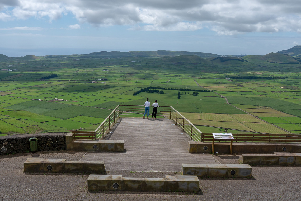 Visitors overlooking the geometric patchwork of green agricultural fields from Serra do Cume Viewpoint on Terceira Island, with volcanic ridges and the Atlantic visible in the distance.
