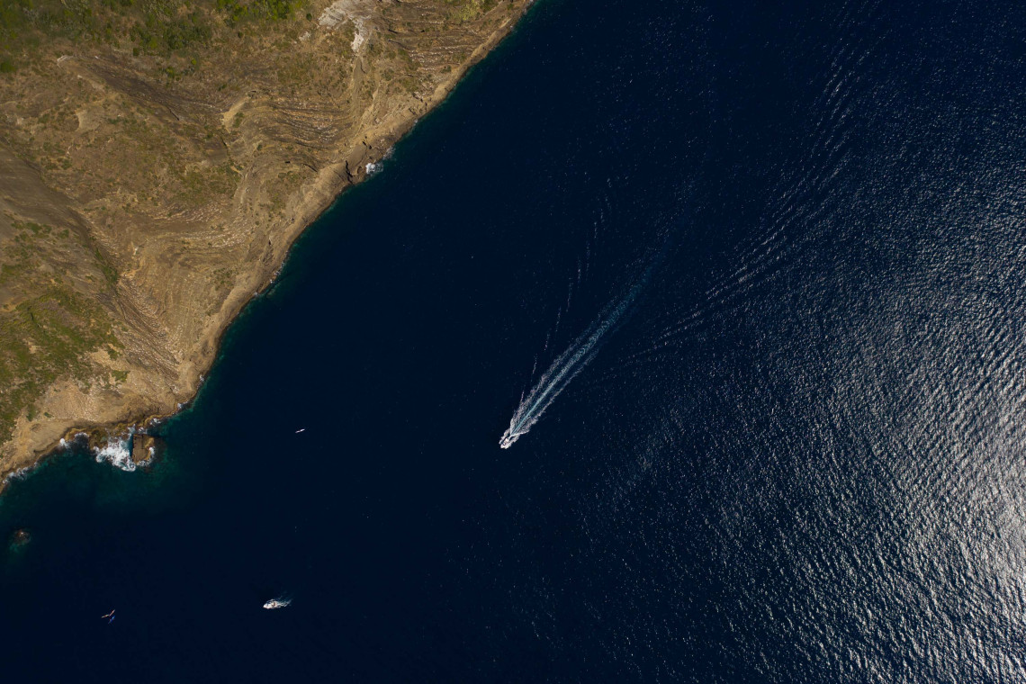 Aerial view of a boat traveling along the rugged volcanic coast of Terceira Island, leaving a white wake across the dark blue Atlantic Ocean.