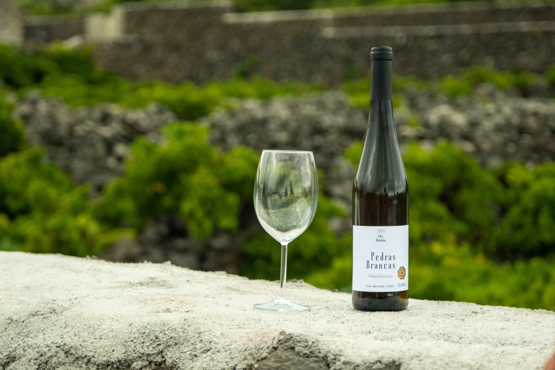 Bottle of Pedras Brancas white wine with a wine glass set against the volcanic-stone landscape of Graciosa Island. The wine is produced from local Azorean grape varieties and reflects the island&rsquo;s volcanic terroir.