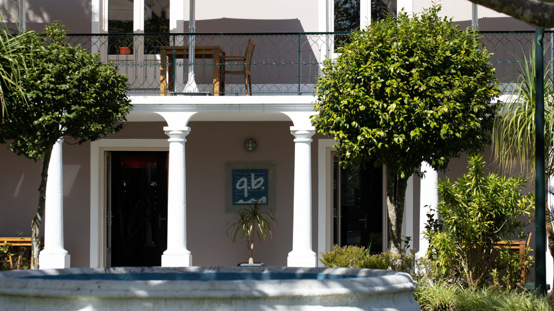 Exterior of Q.B. Restaurante in Angra do Heroísmo, with a covered veranda, white columns and landscaped greenery surrounding the entrance. The restaurant specializes in Portuguese cuisine with regional Azorean products.
