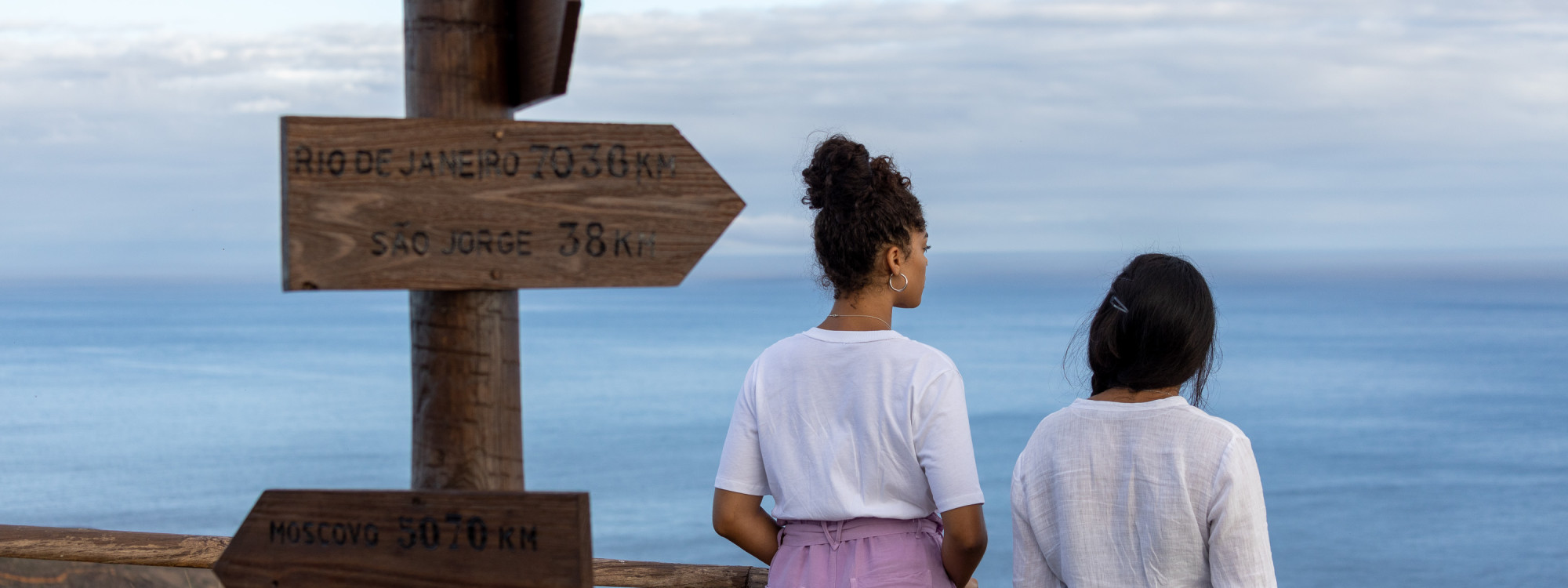 Travelers looking across the Atlantic from Ponta da Restinga Viewpoint on Graciosa Island beside a wooden signpost marking distances to destinations around the world.