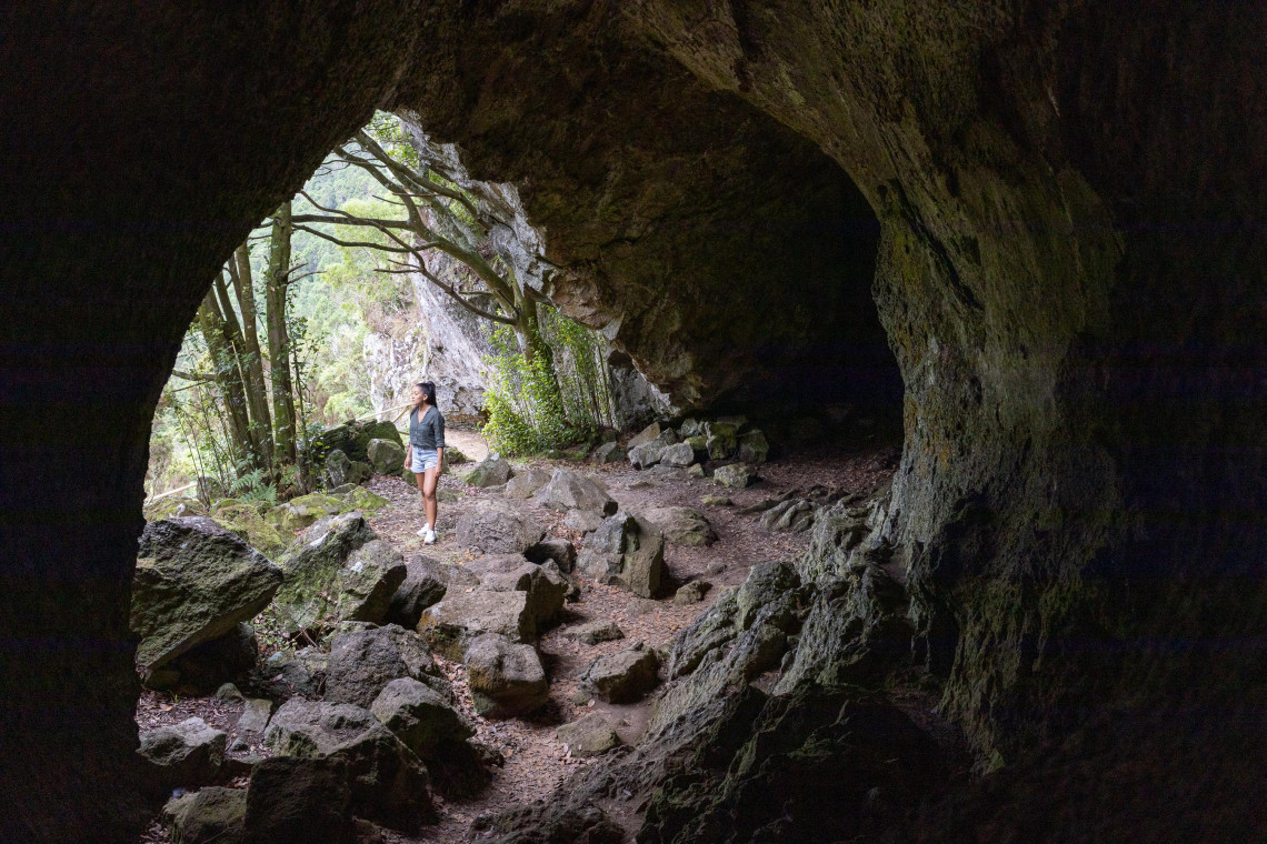 Traveler standing inside Furna da Maria Encantada on Graciosa Island, framed by the dark volcanic cave entrance and surrounding woodland.