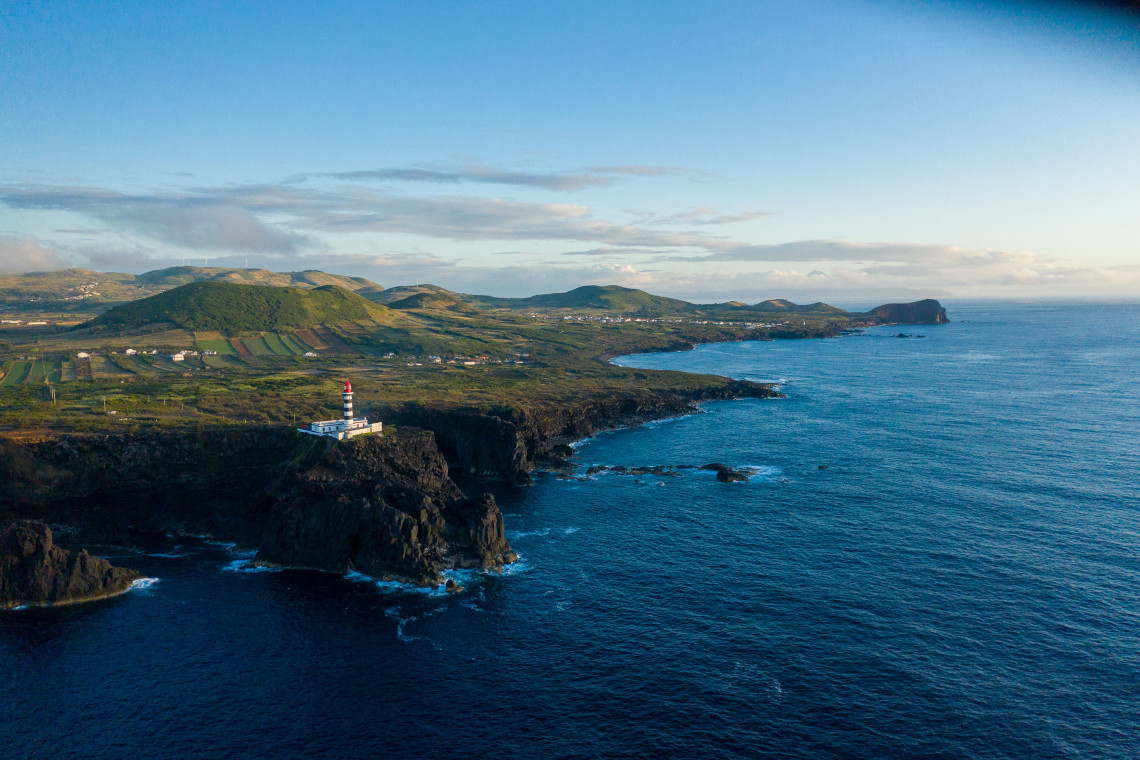 Aerial view of Ponta da Barca Lighthouse on Graciosa Island, standing above rugged volcanic cliffs and agricultural land along the Atlantic coast.