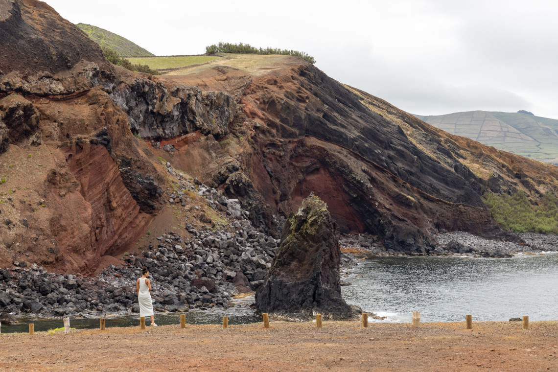 Traveler overlooking Porto Afonso on Graciosa Island, where layered red and dark volcanic cliffs surround a small Atlantic cove and prominent rock formation.