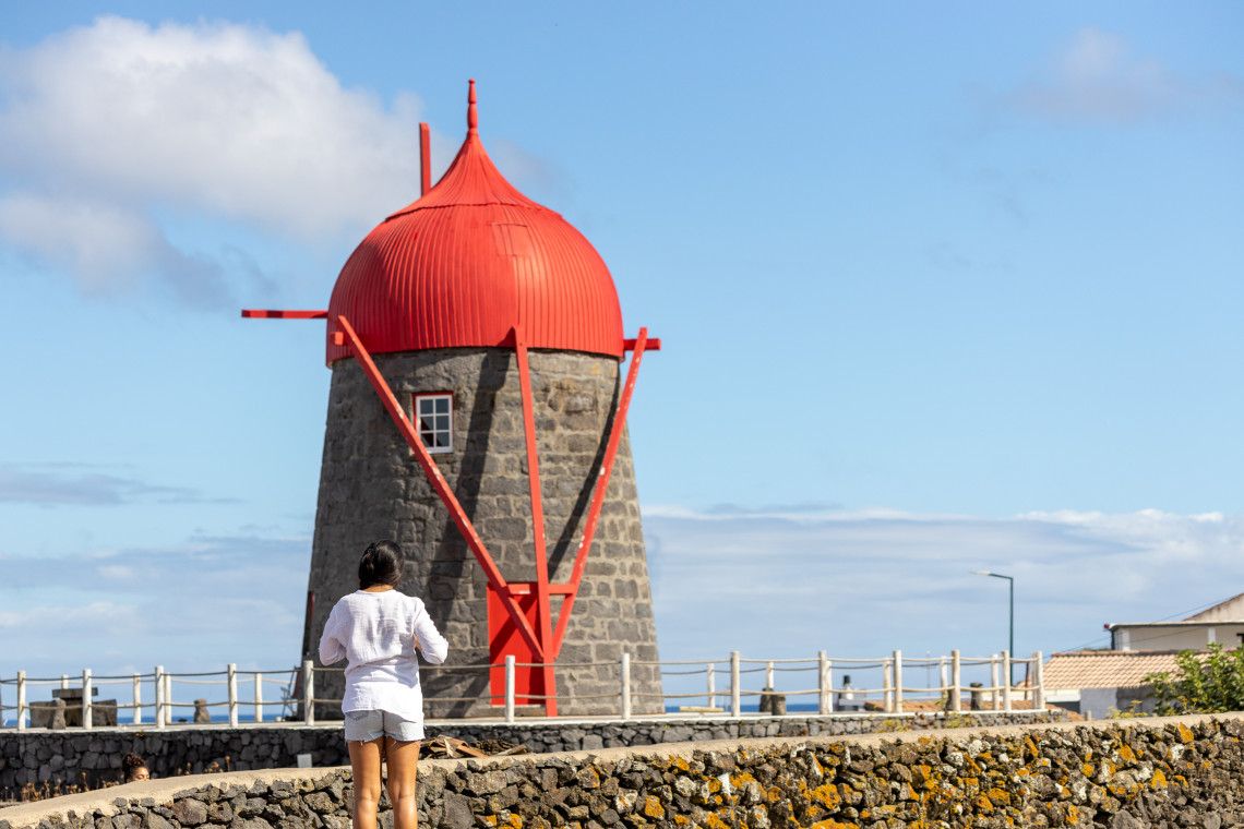Traveler standing beside a traditional basalt windmill in S&atilde;o Mateus on Graciosa Island, distinguished by its large red wooden cap and supports.