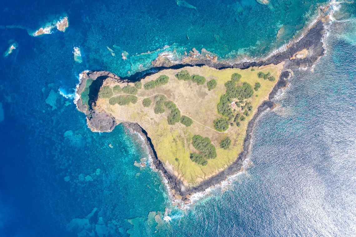 Top-down aerial view of Ilh&eacute;u de Baixo off the southeastern coast of Graciosa Island, showing its grass-covered volcanic surface surrounded by clear Atlantic water.