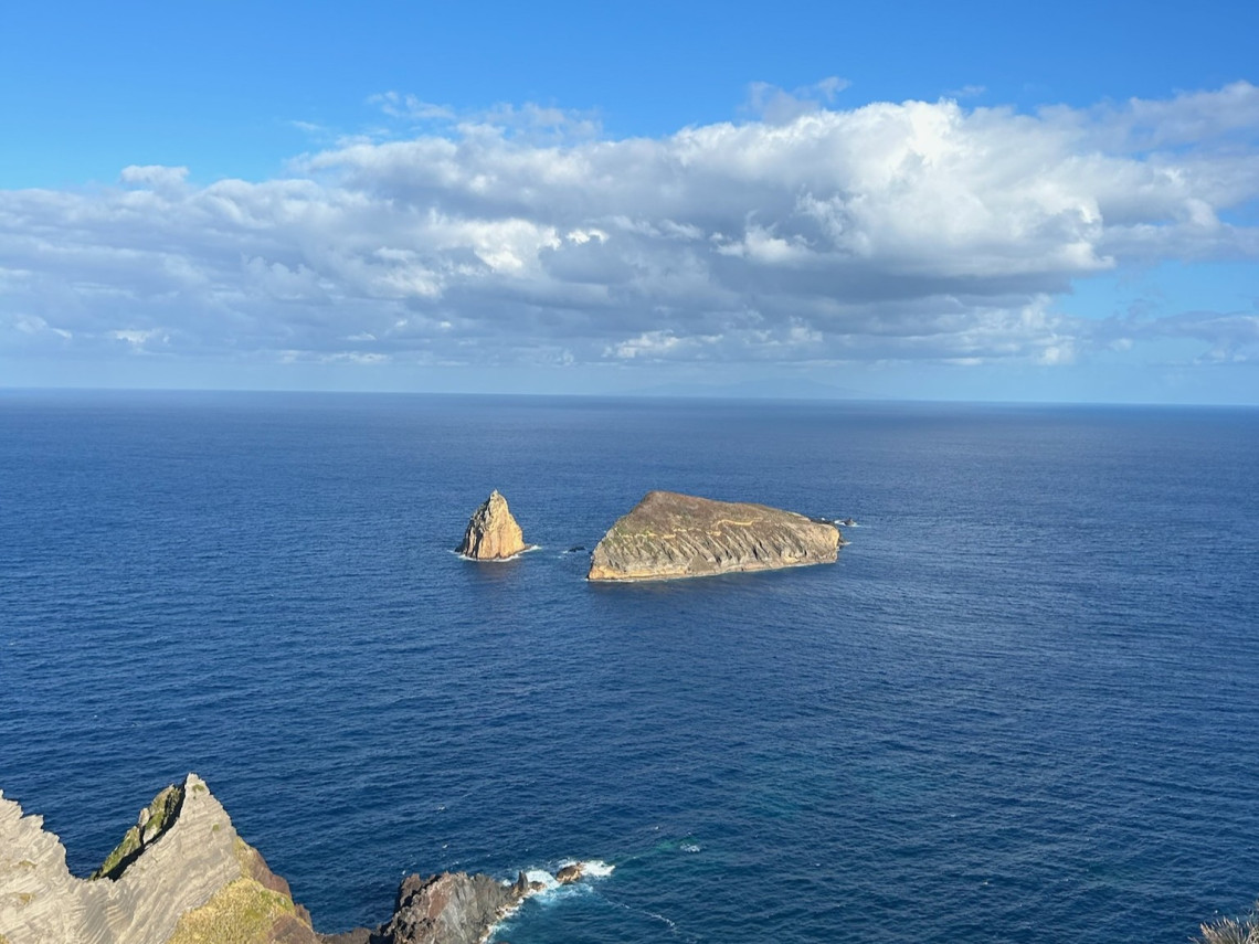 View from Ponta da Restinga on Graciosa Island looking out over the Atlantic Ocean toward two rocky offshore islets. The coastal viewpoint in Luz, Graciosa, shows deep blue water, rugged volcanic cliffs, and a broad sky with scattered clouds.