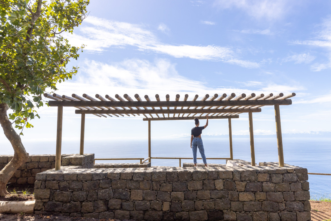 Visitor standing beneath the wooden pergola at Miradouro do Carapacho on Graciosa Island, overlooking the Atlantic Ocean from a basalt stone viewpoint. The scenic overlook in Luz, Graciosa, offers a wide coastal panorama under an open blue sky.